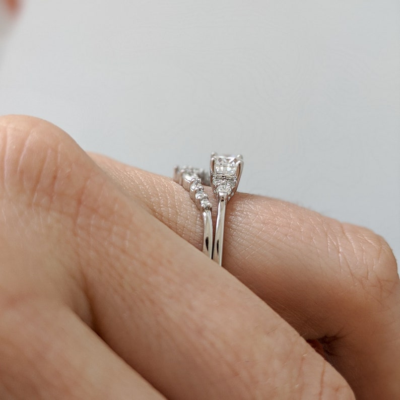 May include: A close-up of a silver ring set with a large, square-cut diamond and smaller diamonds on the band. The ring is worn on a finger.