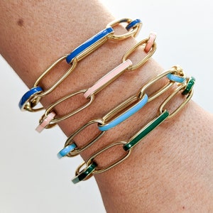 Peut inclure: Quatre bracelets en cha&icirc;ne dor&eacute;e avec des accents en &eacute;mail color&eacute;. Les bracelets sont empil&eacute;s sur le poignet d'une personne. Les couleurs de l'&eacute;mail sont le bleu, le rose, le bleu clair et le vert.