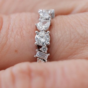 May include: A silver band ring featuring a row of different sized diamonds. The diamonds are round, pear-shaped, and baguette-shaped.