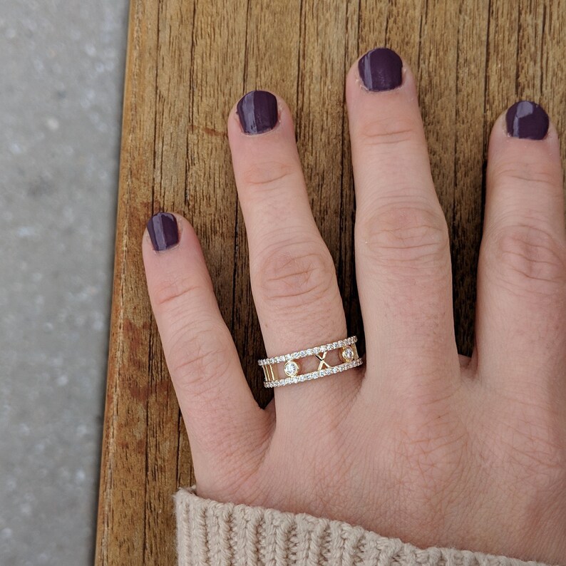 May include: A gold ring featuring a row of small diamonds and Roman numerals. The ring is worn on a person's finger.