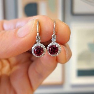 May include: A pair of silver dangle earrings with a red gemstone surrounded by a halo of smaller white gemstones.