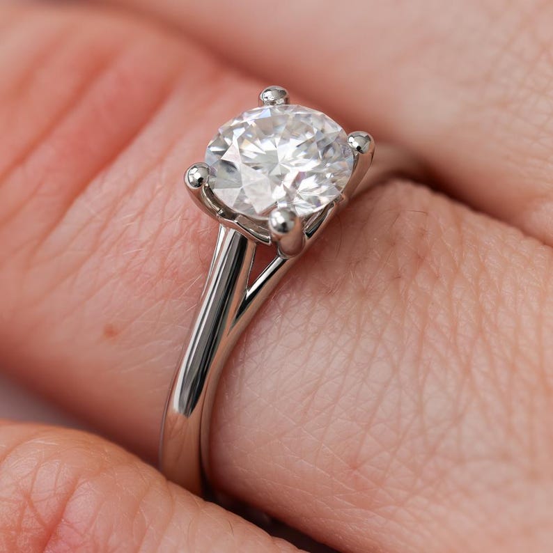 May include: A close-up of a solitaire engagement ring. The ring features a large, round, clear gemstone set in a four-prong setting on a polished silver band. The ring is on a finger.