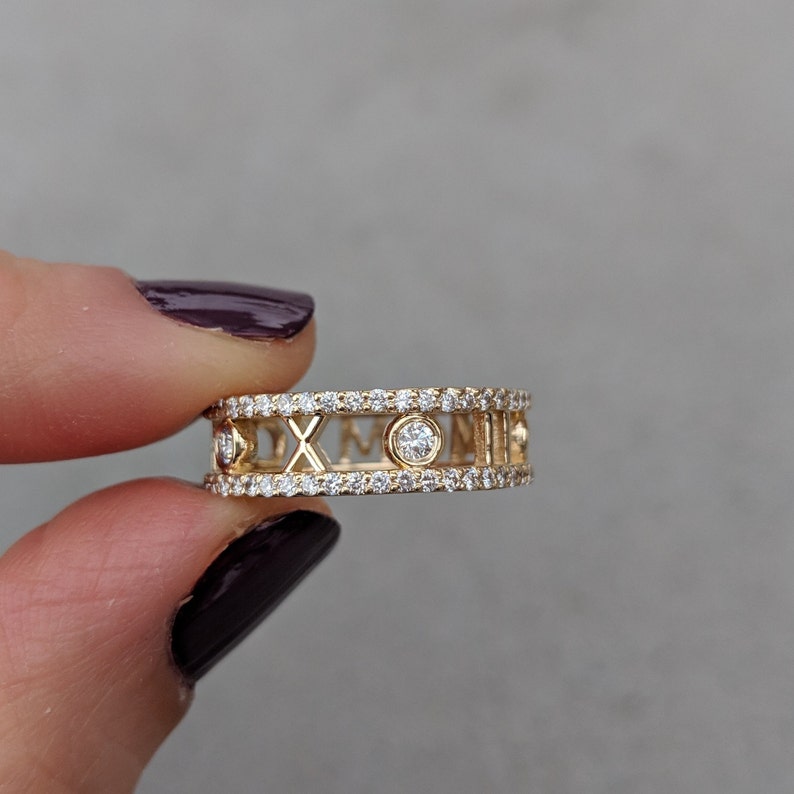 May include: A gold ring with a double row of pave diamonds and Roman numeral accents. A single round diamond is set in the centre of the ring.
