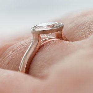 May include: A rose gold engagement ring featuring a bezel-set oval diamond. The ring has a distinctive, open-band design.