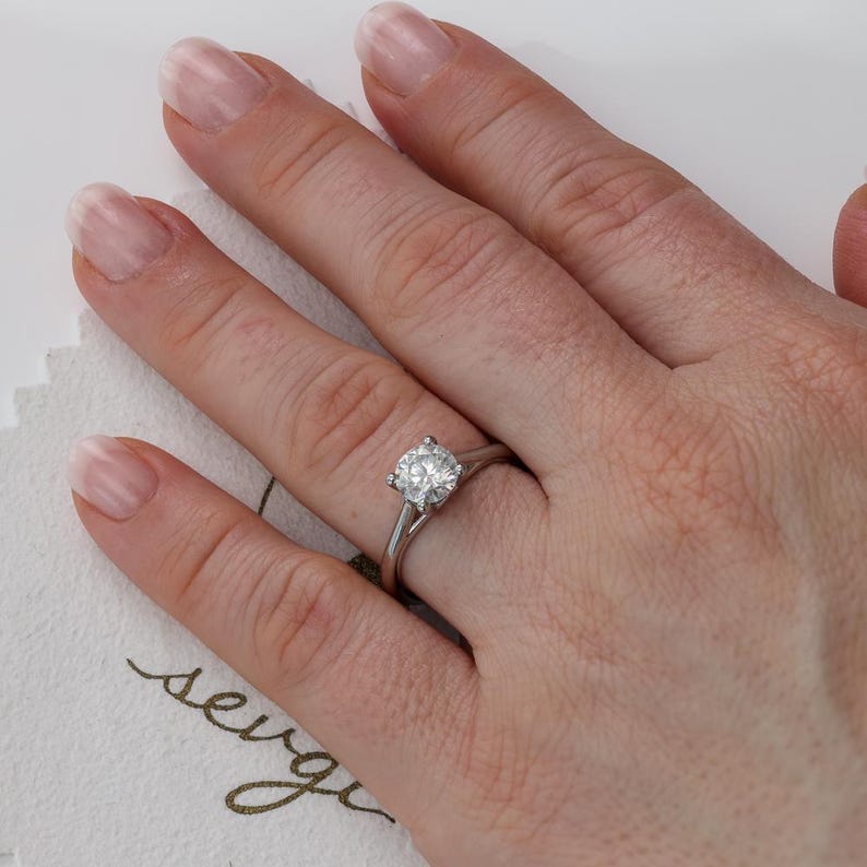 May include: A silver engagement ring with a large, round, clear diamond set in a four-prong setting. The ring is on a person's finger. The ring is on a white background.