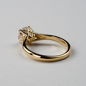 May include: A gold solitaire engagement ring with a large, round, clear gemstone. The ring's band is smooth and polished, and the gemstone is held in place by four prongs. The ring is set against a light background.