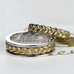 Puede incluir: Dos alianzas de oro con un diseño trenzado. La banda superior es una banda simple, mientras que la banda inferior tiene una banda de metal blanco alrededor del exterior. La inscripción "A&E" está grabada en el interior de la banda inferior.