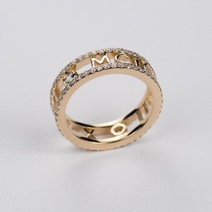 May include: A gold ring with a double band design. The outer band is set with small diamonds and features the letters MCM in a cutout design. The inner band is a solid gold band with a cutout design.