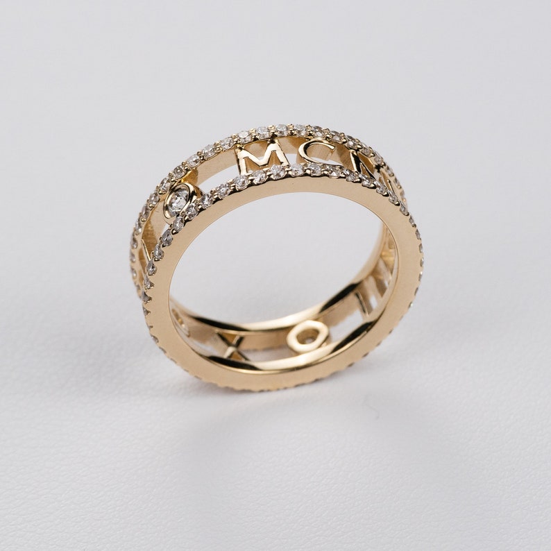 May include: A gold ring with a double band design. The outer band is set with small diamonds and features the letters MCM in a cutout design. The inner band is a solid gold band with a cutout design.