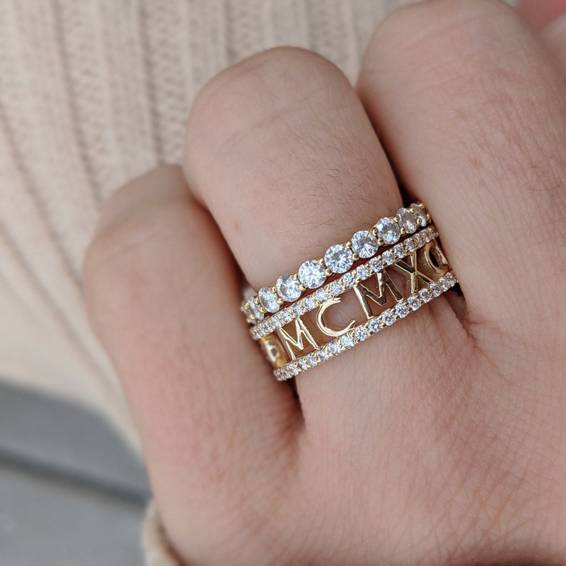 May include: A gold ring with three bands. The top band features the Roman numerals MCMXC. The middle and bottom bands are set with small, clear stones.