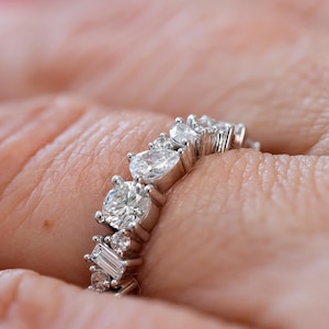 May include: A silver band ring featuring a row of sparkling diamonds. The ring is set with a variety of diamond shapes, including round, oval, and baguette.
