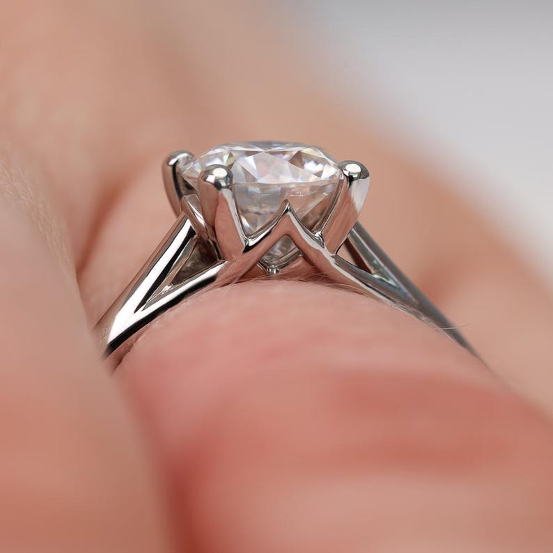 May include: Close-up of a silver engagement ring with a large, clear, round-cut diamond. The ring's band is thin and polished, with a four-prong setting holding the diamond securely. The background is a soft, blurred peach color.