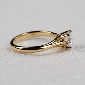 May include: A gold engagement ring with a large, clear gemstone. The ring has a classic four-prong setting and a smooth, polished band. The gemstone is round and sparkles brightly. The ring is set against a neutral background.