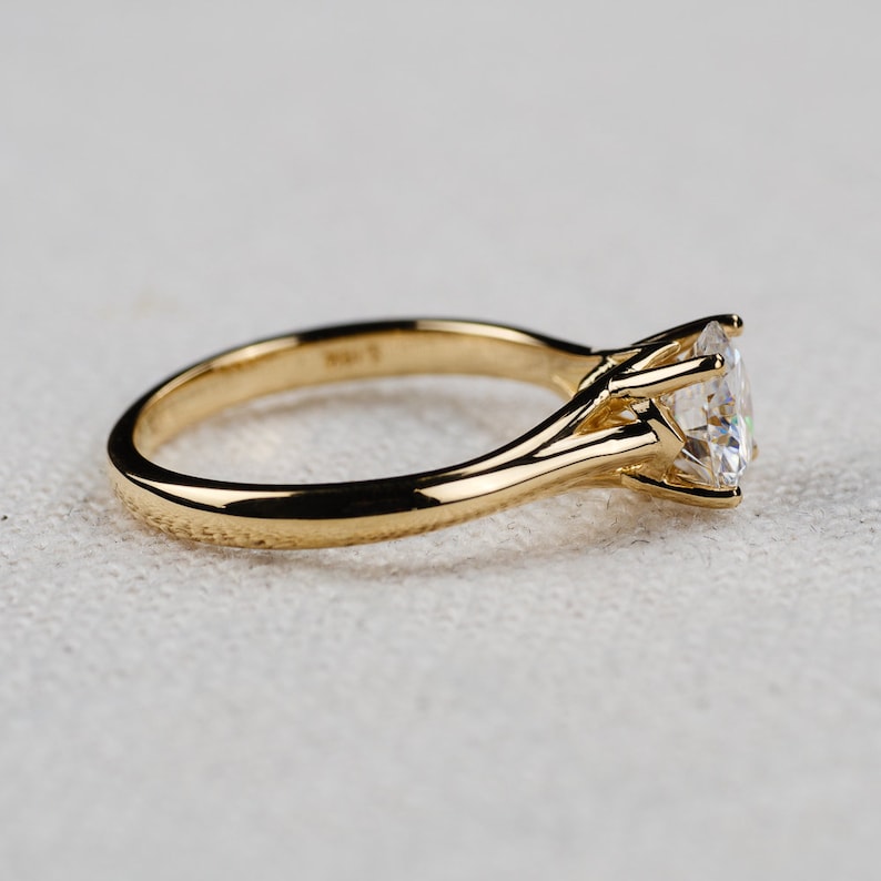 May include: A gold engagement ring with a large, clear gemstone. The ring has a classic four-prong setting and a smooth, polished band. The gemstone is round and sparkles brightly. The ring is set against a neutral background.