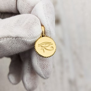May include: Gold pendant featuring an engraved Eye of Horus symbol. The pendant is round and has a beaded edge.