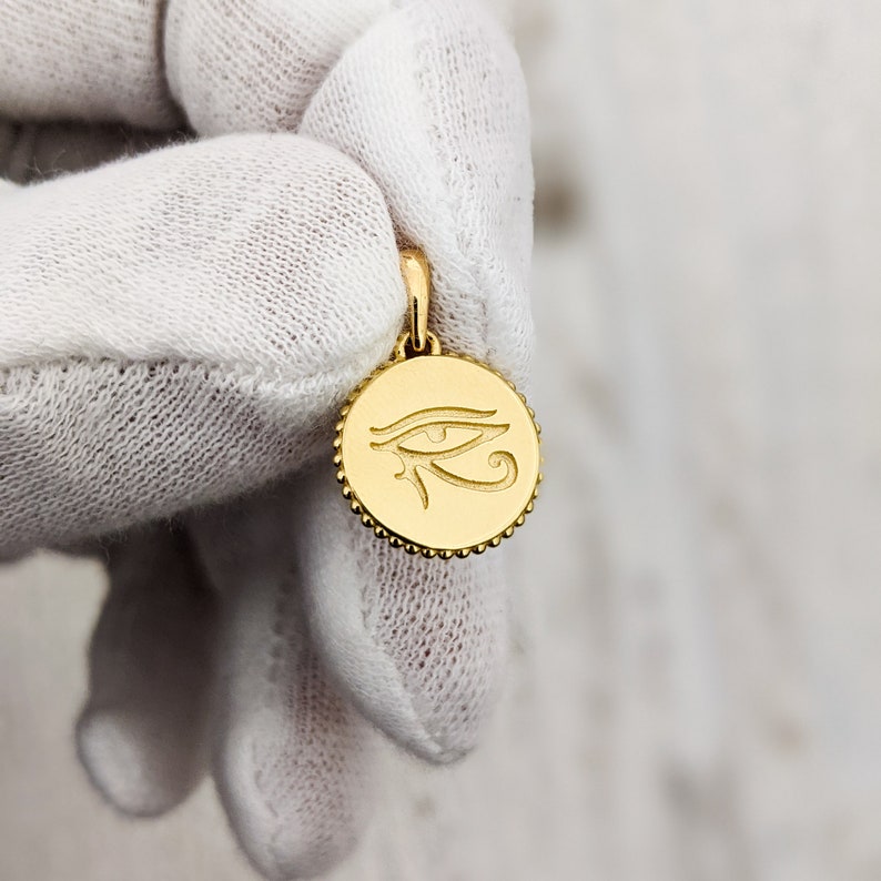 May include: Gold pendant featuring an engraved Eye of Horus symbol. The pendant is round and has a beaded edge.