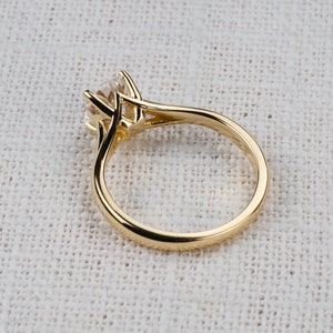 May include: A gold ring with a large, round, clear gemstone. The ring has a classic, elegant design with a four-prong setting. The band is smooth and polished, reflecting the light. The ring is set against a neutral background.