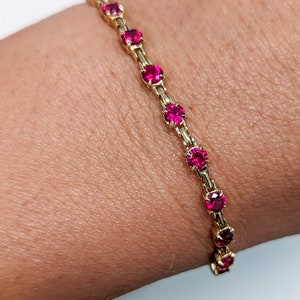 May include: A gold bracelet with a chain design and red gemstones. The bracelet is on a person's wrist.