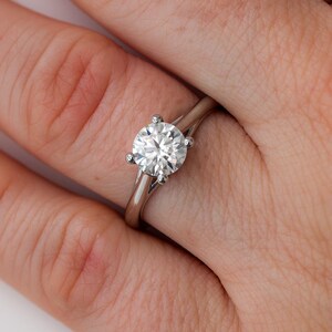 May include: A silver engagement ring with a large, round, clear gemstone. The ring has a simple band and four prongs holding the gemstone. The ring is on a person's finger.