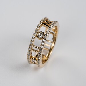 May include: A gold ring with a diamond-encrusted band and a central design featuring the letter "M" in a cage-like setting.