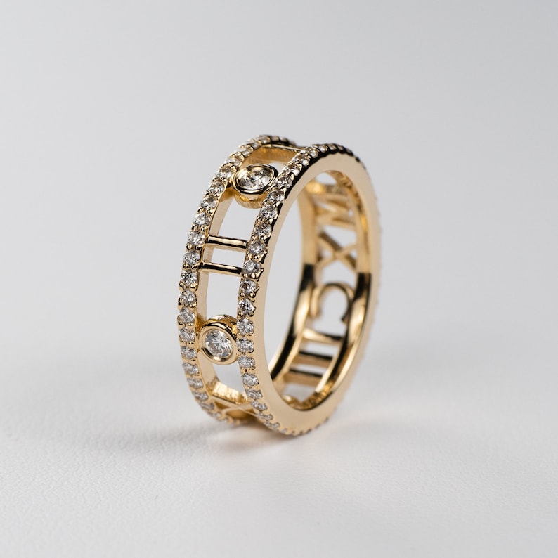 May include: A gold band ring with a row of small diamonds and the Roman numeral 'XII' on the side. The ring has a design of a circle with a line through the center.