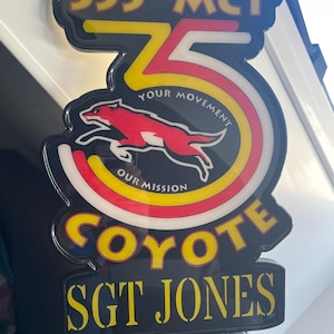 Puede incluir: Un letrero negro y amarillo con el texto "355th MCT" y "COYOTE SGT JONES". El letrero presenta un gráfico de coyote rojo y blanco y las palabras "YOUR MOVEMENT OUR MISSION".