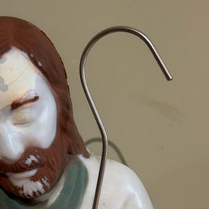 May include: A close-up of a religious figurine with a white robe, red hair, and a brown beard. A silver metal shepherd's crook is in the foreground, curving upwards. The figurine has a serene expression.