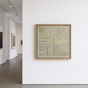 Könnte beinhalten: Ein gerahmtes Kunstwerk mit einem geometrischen Design. Das Kunstwerk zeigt ein quadratisches Muster aus gelben, blauen und weißen Fäden. Der Rahmen besteht aus hellem Holz, und das Kunstwerk wird in einer Galerie an einer weißen Wand ausgestellt.