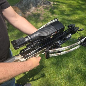 Op de afbeelding: Een zwarte en camouflage kruisboog met een vizier en de tekst "Oracle X". De kruisboog wordt door een persoon op een grasachtige achtergrond vastgehouden. De merknaam "TenPoint" is zichtbaar.