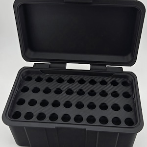 Könnte beinhalten: Schwarze rechteckige Aufbewahrungsbox mit offenem Deckel. Der Innenraum hat einen Einsatz in Carbonfaseroptik mit mehreren kreisförmigen Aussparungen. Die Box hat einen sicheren Verschluss.