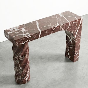 May include: A contemporary console table crafted from red marble with white veining. The table has a rectangular top supported by a twisted column and a squared arch. The table measures approximately 122 cm long, 36 cm wide, and 76 cm high.