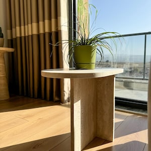 Puede incluir: Una pequeña mesa auxiliar de travertino beige con una tapa redonda y una sola pata rectangular. Una planta en maceta verde con hojas largas y finas está sobre la mesa. La mesa está frente a una ventana.