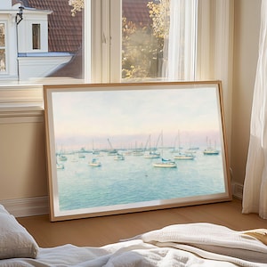 May include: A framed art print depicting sailboats on a tranquil, blue sea. The artwork, in a watercolour style, is set within a light wood frame. It leans against a wall near a window, evoking a calm, nautical atmosphere.