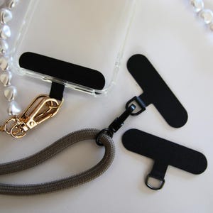 Peut inclure: Un étui de téléphone transparent avec une languette noire et un fermoir doré attaché à une sangle en corde grise. Deux languettes noires supplémentaires avec des boucles sont également visibles. Un collier de perles est en arrière-plan.