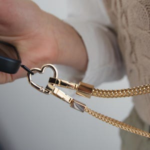 Op de afbeelding: Goudkleurige telefoonriem met een hartvormige sluiting. De riem heeft een geweven kettingontwerp en twee rechthoekige sluitingen. De telefoonhoes is zwart. De achtergrond is een persoon die een wit overhemd en een beige trui draagt.