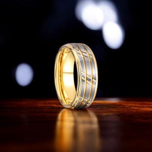 Puede incluir: Un anillo de oro y plata con un diseño geométrico. El anillo tiene un interior dorado y un exterior plateado con una serie de líneas paralelas y acentos diagonales. El anillo se muestra sobre una superficie de madera oscura.