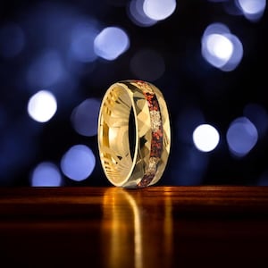 May include: A gold ring with a geometric design and a central band of red and gold stones. The ring is set against a blurred background with bokeh lights, creating a luxurious feel.