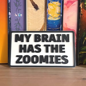 May include: A white rectangular sign with a black border and the phrase "MY BRAIN HAS THE ZOOMIES" in bold, black letters. The sign is displayed in front of a row of colourful book spines.