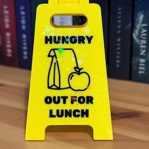 May include: A bright yellow plastic sign with the words "HUNGRY" and "OUT FOR LUNCH" in black. The sign features a graphic of a lunch bag and an apple. The sign is standing upright on a wooden surface.
