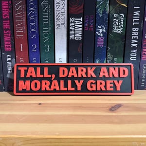May include: A rectangular sign with the text "TALL, DARK AND MORALLY GREY" in red, raised lettering on a black background. The sign is displayed in front of a shelf of books.