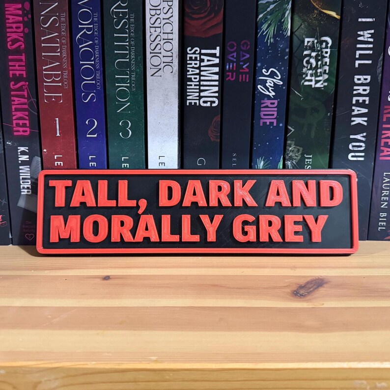 May include: A rectangular sign with the text "TALL, DARK AND MORALLY GREY" in red, raised lettering on a black background. The sign is displayed in front of a shelf of books.