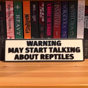 May include: A white sign with black text stating "WARNING MAY START TALKING ABOUT REPTILES" is placed in front of a bookshelf. Various books with colourful spines are visible, including titles such as "Heavy" and "Voracious".