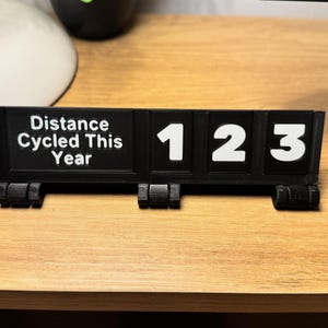 May include: A black 3D-printed cycling distance counter with white numbers and text. The text reads "Distance Cycled This Year". The counter displays the numbers 123. The item is on a wooden surface.