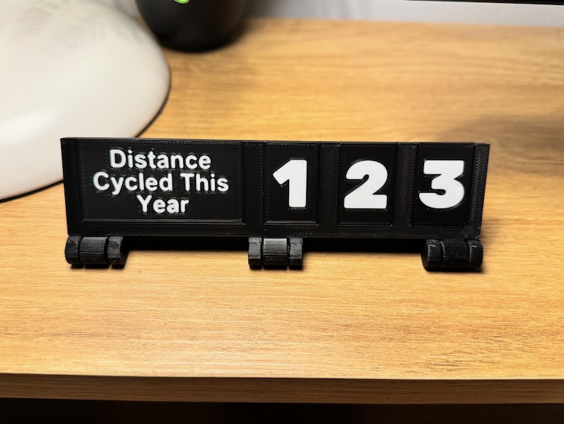 May include: A black 3D-printed cycling distance counter with white numbers and text. The text reads "Distance Cycled This Year". The counter displays the numbers 123. The item is on a wooden surface.