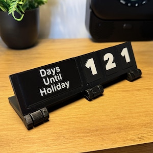 Könnte beinhalten: Ein schwarzer Schreibtischkalender mit weißen Zahlen und dem Text "Days Until Holiday". Der Kalender zeigt "121" verbleibende Tage an. Der Kalender besteht aus schwarzem Kunststoff und steht auf einer Holzoberfläche.