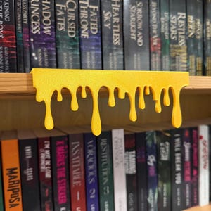 May include: A yellow, dripping-style shelf decor piece. The shelf sits above a row of books with various titles. The decor has a flat top and a series of downward-flowing drips, adding a unique touch to a bookshelf.