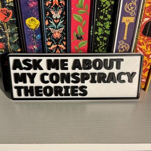 May include: A rectangular sign with the text "ASK ME ABOUT MY CONSPIRACY THEORIES" in black, raised lettering. The sign has a white background and a black border. Decorative book spines are visible in the background.