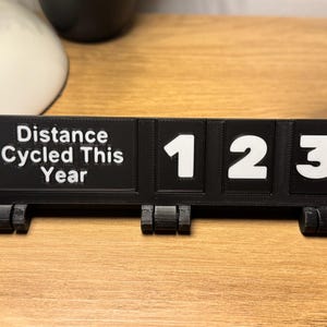May include: A black 3D-printed distance tracker with white numbers and text. The text reads "Distance Cycled This Year." The numbers displayed are "123". The tracker is on a wooden surface.
