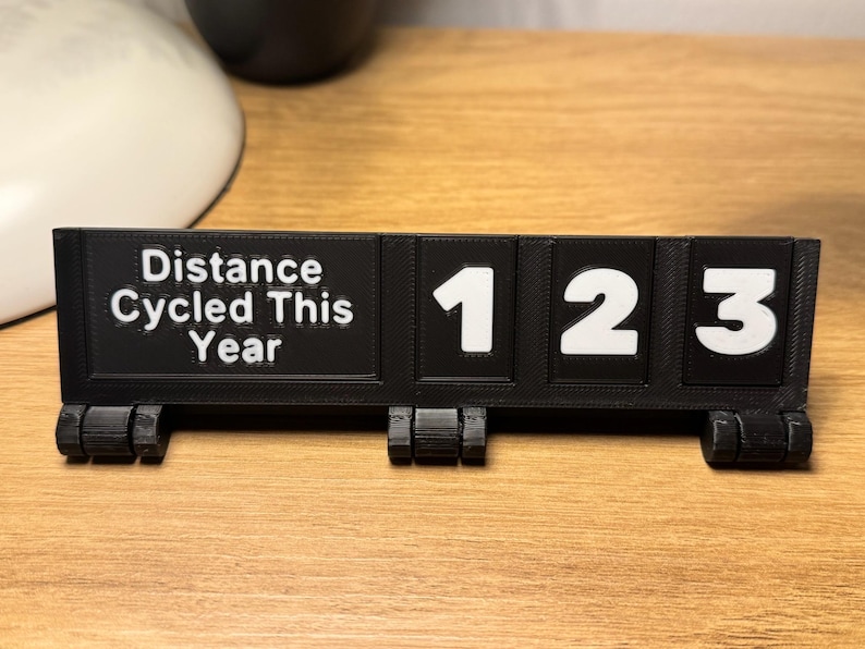 May include: A black 3D-printed distance tracker with white numbers and text. The text reads "Distance Cycled This Year." The numbers displayed are "123". The tracker is on a wooden surface.