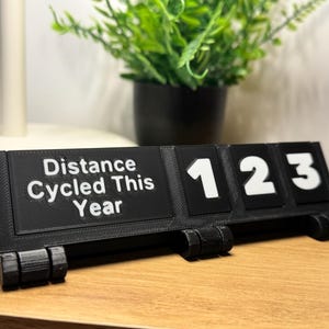 May include: A black desk display with the text "Distance Cycled This Year" and the numbers "123" in white. The display is made of a matte black material and sits on a wooden surface. A small potted plant is in the background.